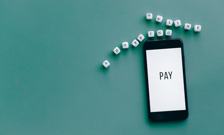 Smartphone com a palavra “PAY” cercado por dados com a expressão “online shopping”, sobre fundo azul-esverdeado.