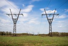 Linhas de transmissão de energia atravessam um campo verde sob céu azul com poucas nuvens.