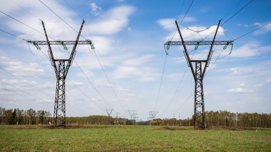 Linhas de transmissão de energia atravessam um campo verde sob céu azul com poucas nuvens.