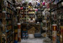 Lojista atrás do balcão de uma pequena loja com diversos produtos expostos nas prateleiras.