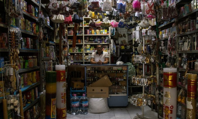 Lojista atrás do balcão de uma pequena loja com diversos produtos expostos nas prateleiras.