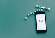 Smartphone exibindo a palavra “PAY”, cercado por dados com letras sobre fundo azul-turquesa.